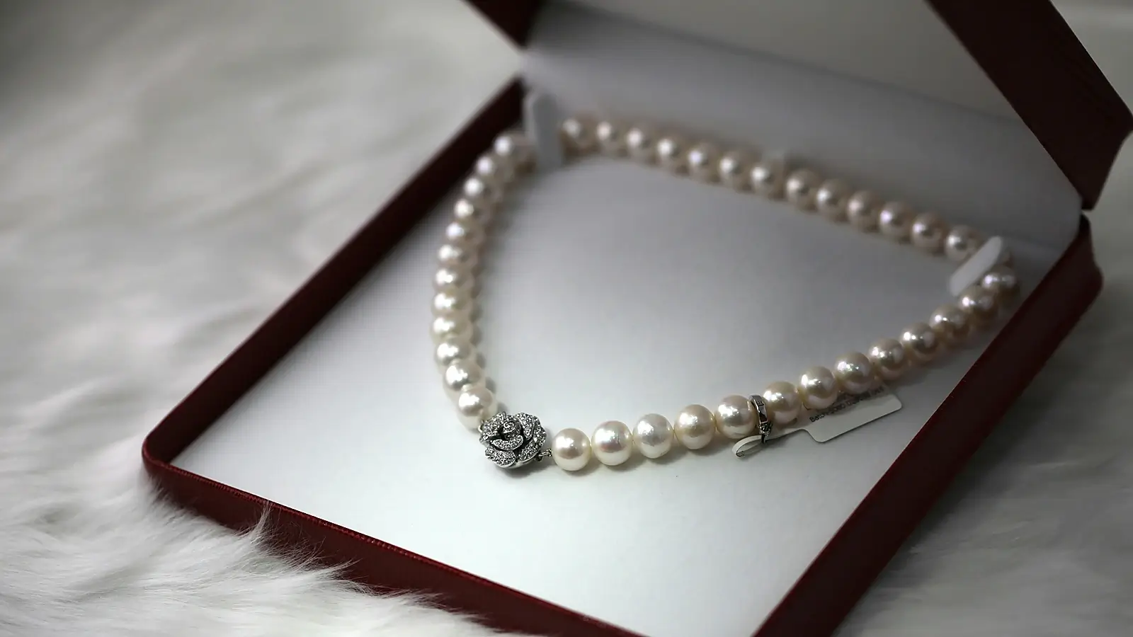 Elegant pearl necklace with a decorative jeweled clasp displayed inside an open jewelry box on a soft white surface.